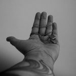 Black and white photo of a tattooed arm holding a pendant. Artistic and moody composition.