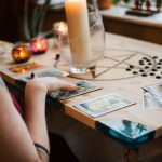 Crop unrecognizable female fortune teller predicting fate with tarot cards near burning candles on table at home